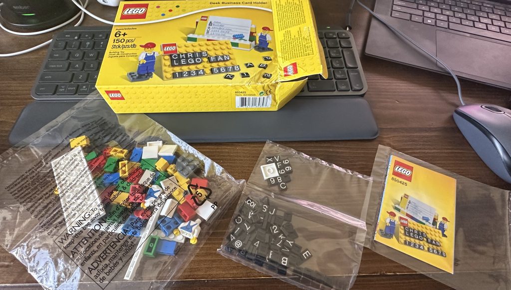 Photo of opened LEGO set 850425 Desk Business Card Holder showing instruction manual, lettered tiles, and random brick in three separate bags