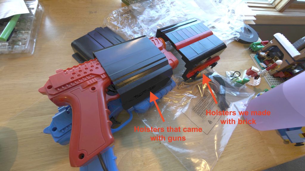 Arcade light guns shown sheathed inside a black plastic bracket that came with the guns. Just in front of it, new brackets that are made out of brick instead.